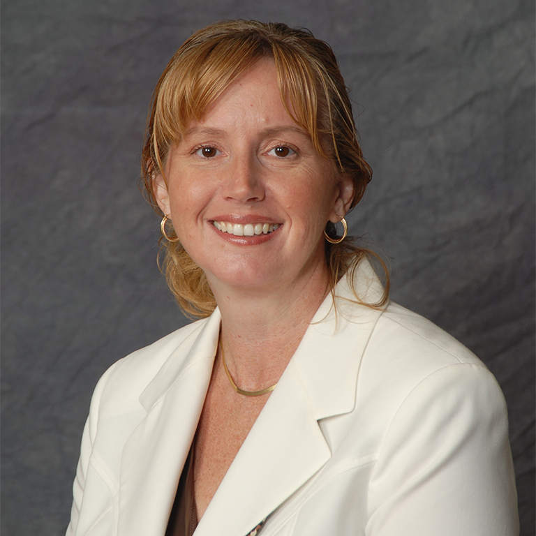 Elizabeth Wager sits in front of a gray background wearing a white blazer.